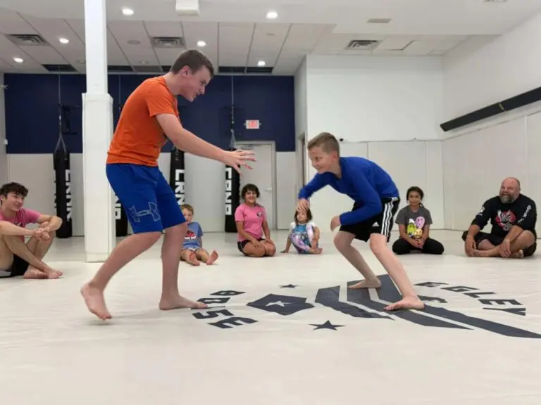 Young students sparring in a Brazilian Jiu Jitsu class while teammates watch from the sidelines.